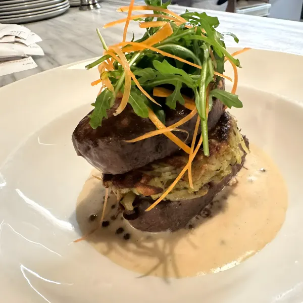 A tall stack of grilled steak on a potato cake with rocket and a creamy sauce